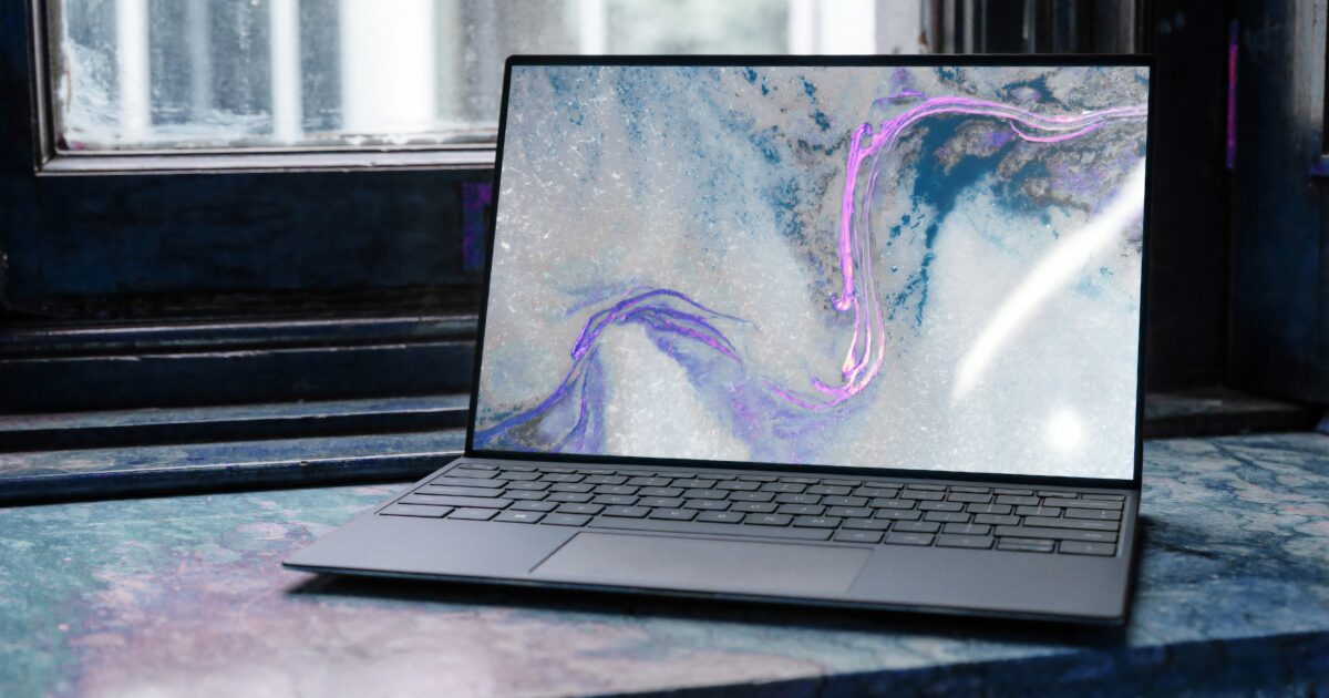 窓際のカラフルなテーブルに置かれたノートパソコンに抽象的なアートの壁紙が表示されている様子