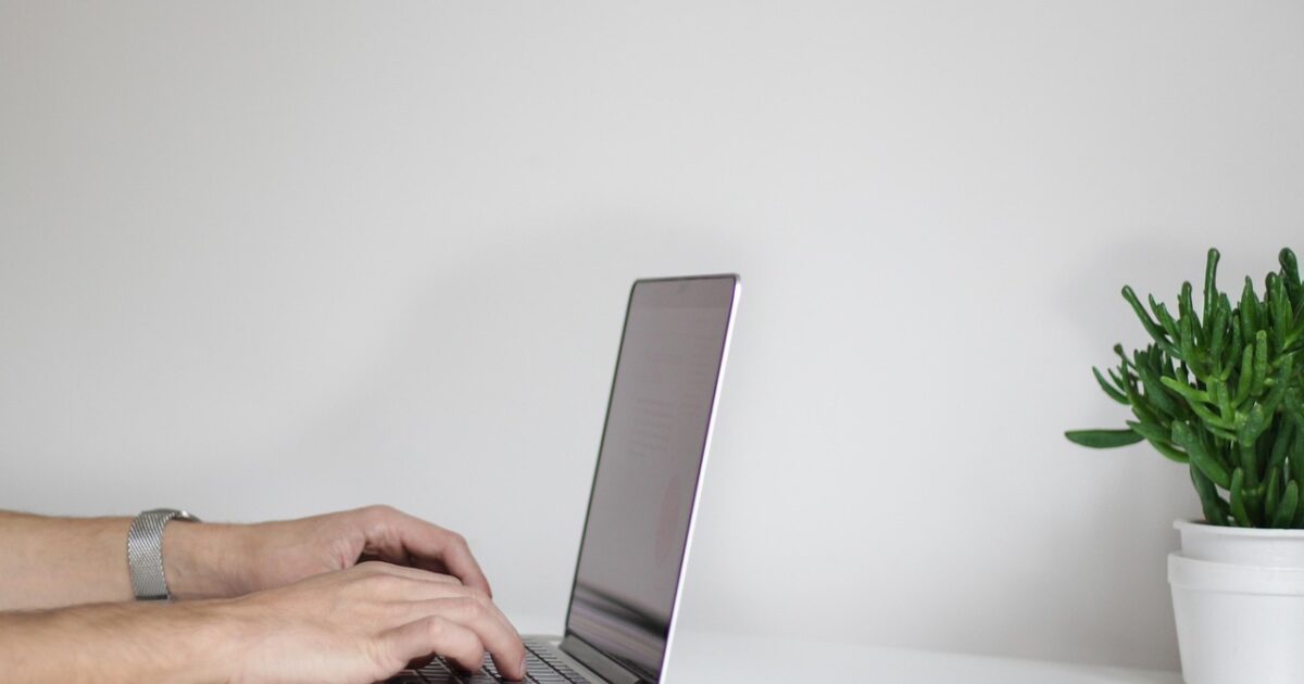 白いデスクでノートパソコンを操作している手元と観葉植物のシンプルな作業スペース