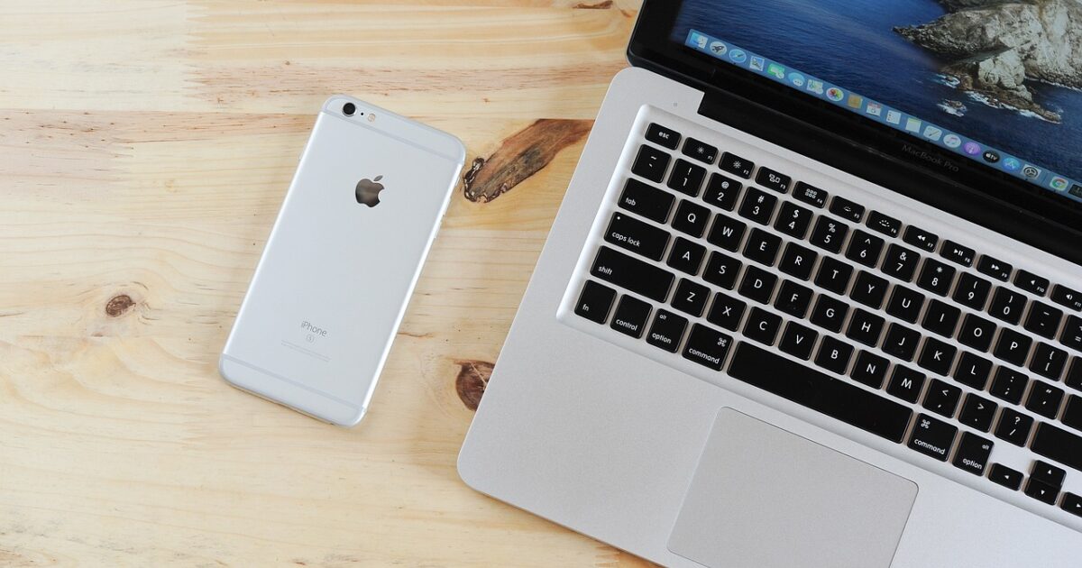 木製デスクの上に置かれたMacBook ProとシルバーのiPhoneのクローズアップ