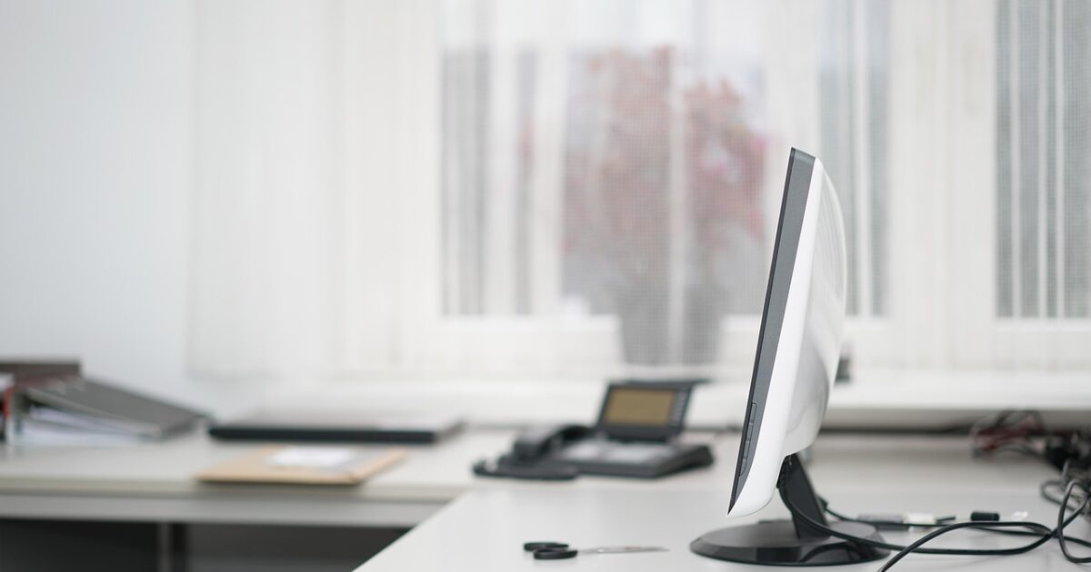 白いカーテンの前にあるオフィスデスクにモニターと書類が置かれたシンプルな作業環境